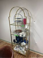 Front view of a tall metal and glass display rack, gold frame with arch top, multiple shelves with glass and ceramic items (not included).