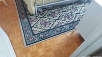 Top-down photo of floral patterned area rug partially under a bed on parquet flooring showing border and some wear.