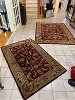 Two wool rugs laid on a tiled floor, showing both rugs with floral vine patterns in red and green tones.