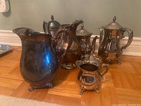 Front view of silver-plated pitchers, teapots, sugar bowls, and creamers grouped together on floor.