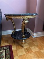Side view of ornate two tier black and gold side table with black marble effect resin tops next to wall corner and hardwood floor