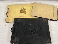 Three photo albums on a white surface, two lighter cream colored and one black with embossed covers