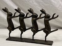 Front view of the sculpture showing four rabbits dancing in a row with raised arms and one leg, mounted on a rectangular base.