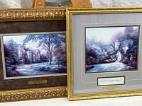 Two framed Thomas Kinkade prints shown side by side, one with an ornate gold frame, the other with a simpler gold frame, with plaques visible under each print