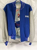 Front view of blue and gray souvenir jacket with name 'Phil' on chest and partially visible inside satin lining embroidery.