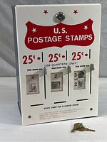 Front view of vintage U.S. postage stamp vending box showing three 25¢ coin slots and red U.S. Postage Stamps text with lock and keys at base.