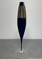 Full view of the tall art glass vase standing on a grey surface against a white wall, showing its overall shape and proportions.
