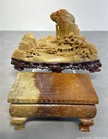 Close-up of soapstone carving showing intricate details of houses, trees, and rocky mountain mounted on a carved wooden base. Positioned behind is the carved wooden stand.