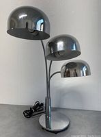 Chrome table lamp showing all three domes facing forward, lamp base, and power cord laid out on a flat surface.
