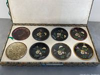 Eight round sample cloisonne plates displayed inside box lid showing detailed floral designs on dark backgrounds