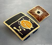 Top view showing the two enamel belt buckles side by side on a grey surface, highlighting their colors and shapes.