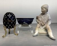 Three items displayed side by side: decorative blue and silver embellished egg music box on gold stand, blue and white Asian style bowl, bisque figurine of seated child holding teddy bear