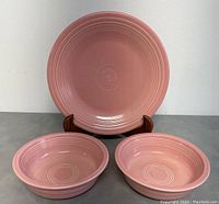 View of the pink 10.5 inch plate displayed on a wooden stand with two matching shallow bowls in front.