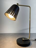 Front angle view of modernist table lamp showing perforated metal shade, brass arm, and round black base on surface
