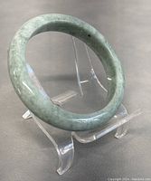 Jade bangle displayed on a clear acrylic stand showing round shape, natural light green color with polished finish and some darker inclusions.