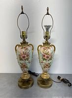 Full view of the pair of urn table lamps side-by-side showing floral artwork and brass bases with electrical cords.