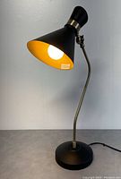 Front view of the retro table lamp illuminated showing black conical shade with golden interior and brass arm.