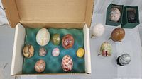 Top-down view of 10 polished egg-shaped stones resting on green felt inside a cardboard box, displaying varied natural patterns and colors including yellow, green, white, brown, and red hues.