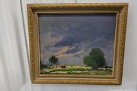 Frontal view of framed oil painting depicting stormy sky over rural landscape with two trees and small buildings