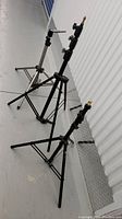 Three black telescopic stands arranged on a floor, varying in height and detailing, one with a ballhead pivot.