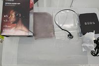 Photo of the Monster Open Ear HP bone conduction headphones in plastic tray, soft carrying pouch, charging cable, and user manual laid out next to packaging box.