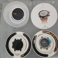 Two Motorola Moto 360 smart watches in round white packaging with multiple wristbands, manuals and charger.
