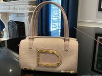 Front view of barrel-shaped lavender fabric and leather handbag with gold-tone D medallion on reflective surface