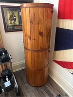 Full front view of the tall wooden barrel standing against a wall, showing natural wood finish and metal hinges.