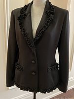 Full front view of black jacket on mannequin showing ruffled collar, flap pockets and hem trim
