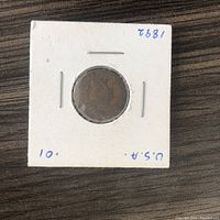 Front side of 1892 Indian Head penny in protective white cardboard holder with handwritten year and country.