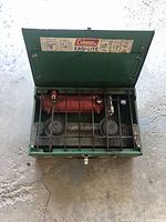 Open green metal Coleman Easi-Lite portable stove showing two burners, fuel tank, and protective grates inside.
