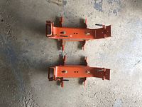 Two orange metal adjustable platform brackets laid on floor, showing overall shape, handles, and adjustment holes.