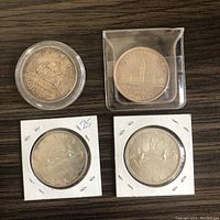 Four Canadian silver dollars placed on a wooden surface, two in plastic holders and two in white cardboard 2x2 holders. Front sides showing the reverse designs: Bluenose schooner and Parliament buildings.