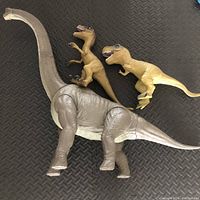 Three dinosaur toys on black textured surface, including a large gray Brachiosaurus with articulated neck and tail and two smaller T rex rubber toys