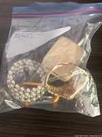 Clear zip-lock bag with assorted jewelry and small items including faux pearl string, gold-tone circular brooch or pendant, key-shaped item, and light stone.