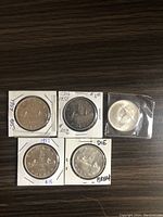 Five silver Canadian dollar coins placed on a wooden surface. Four coins are in cardboard holders with handwritten notes, and one coin in plastic sleeve. Obverses and reverses visible.