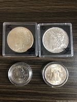 Four US silver coins placed on a wooden surface in clear plastic protective holders, front view showing obverse sides of the Peace Dollar, Morgan Dollar, SBA Dollar, and Silver Half Dollar.