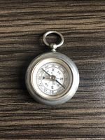 Image of vintage pocket compass showing the front face, circular metal casing, glass covering the white dial with compass needle and directional markings, placed on a wooden surface.