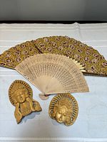 Photo of two carved wooden Asian-style statuette heads and two folding fans on a white fabric surface.