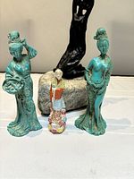Front view of four statuettes showing two turquoise ceramic women, one smaller multicolored figure, and one black abstract sculpture on stone.