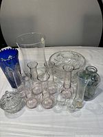 Front view of entire glassware collection showing multiple small clear glass vases, one large clear flared vase, blue etched vase, round bowl, jar with stones, and lidded container.
