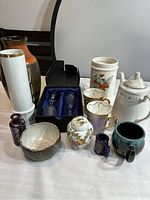 Overview of ceramics, porcelain, pottery items including mugs, teapot, decorative egg, bottles, bowl, and decanters on table.