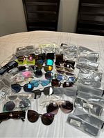 Photo showing a full collection of various styles and colors of new sunglasses with protective packaging on a white table, captured with black chairs in background.