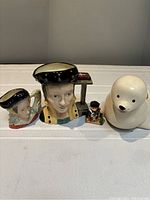 Collection of four porcelain items displayed on a flat surface including two face-shaped pitchers, a small soldier figurine, and a white seal figurine.