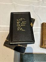 Black hardcover book titled 'Shalte Gottes Gebote' with gold floral design, placed on top of other vintage books