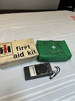 Photo showing closed white metal vintage first aid kit box, closed green plastic Vantage first aid kit box, and Vantage portable radio on a table