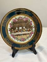 Front view of decorative plate displaying The Last Supper scene, floral accents, and gold rim on stand.