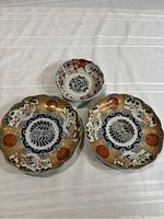 Three vintage Asian Imari plates arranged on a white cloth; two round plates and one smaller scalloped bowl.