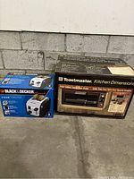 Two boxed kitchen appliances on concrete floor against block wall: Toastmaster under-cabinet toaster oven and Black & Decker 2-slice toaster.