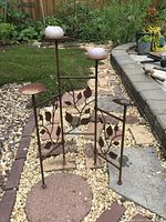 Outdoor rustic metal candle holder with multiple platforms and leaf decorations standing on gravel outdoors.